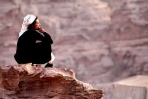 A Jordanian Bedouin sitting on the edge of red rocks in Petra. Photograph.